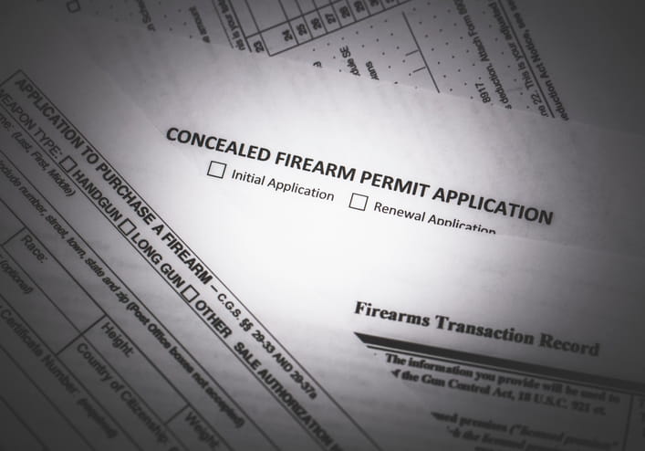 A close-up, top-down view of several official documents, including a "Concealed Firearm Permit Application" with checkboxes for Initial and Renewal applications, and an "Application to Purchase a Firearm.