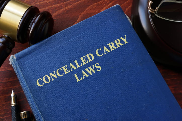 Top-down view of a blue law book titled "Concealed Carry Laws" in gold lettering, placed on a wooden desk next to a judge's gavel and a fountain pen