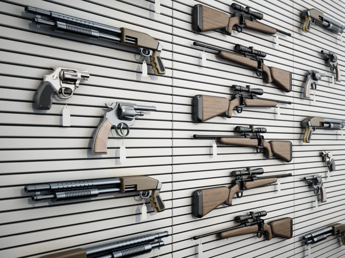 A variety of firearms including rifles with scopes, shotguns, and revolvers displayed on a white slatwall in a gun shop or showroom, each with a small white price tag hanging from it.
