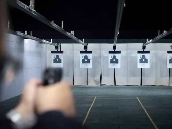 Point-of-view shot of a person aiming a handgun at silhouette paper targets at an indoor shooting range with multiple lanes.