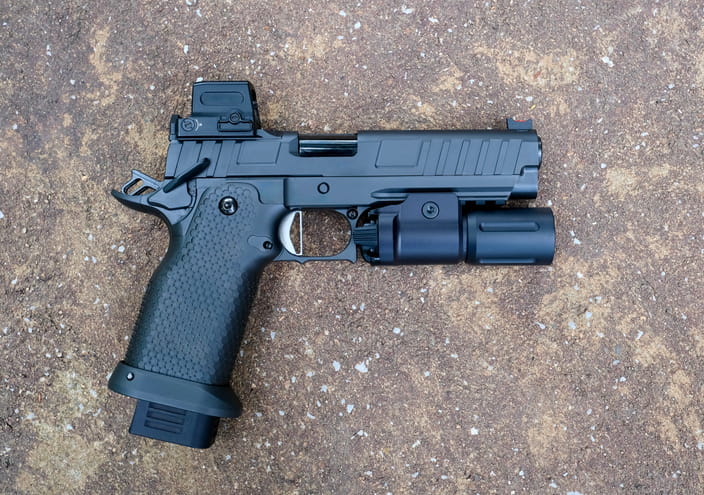 A modern black tactical handgun featuring a red dot optic sight and a mounted flashlight, resting on a textured concrete surface.