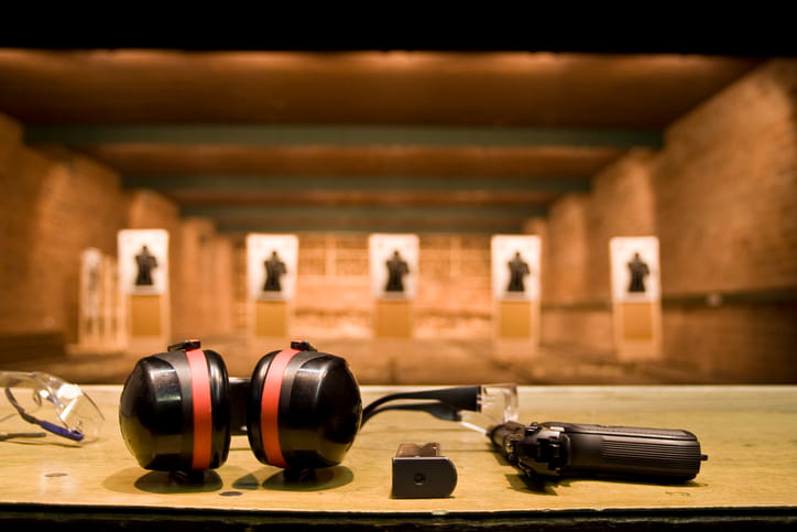 A pair of black and red ear muffs, clear safety glasses, a pistol magazine, and a handgun rest in focus on a wooden counter, with indoor shooting lanes and paper silhouette targets blurred in the warm-lit background.