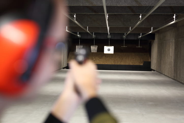 Over-the-shoulder view of a blurred person wearing ear protection and aiming a handgun down an indoor shooting range lane toward a paper target in focus.