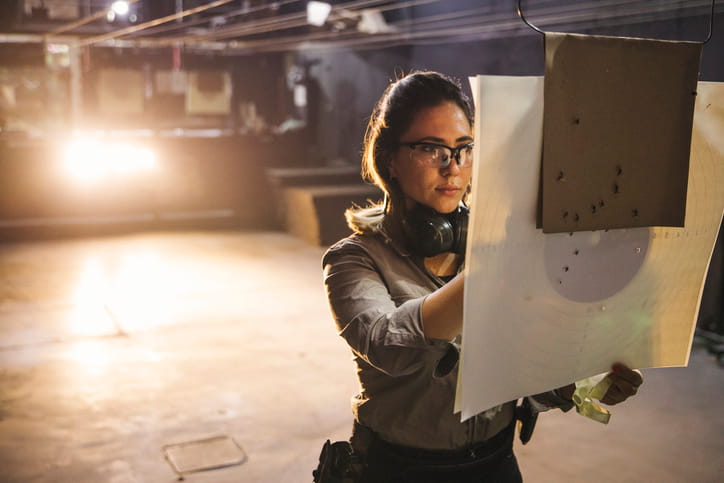 A woman wearing safety glasses and ear protection around her neck examines a paper shooting target with bullet holes at an indoor gun range.