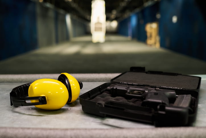 Yellow ear protection headphones next to an open gun case with a handgun, set on a table at an indoor shooting range.